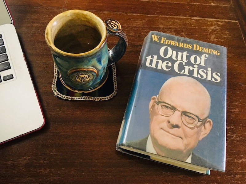 Deming's book Out of the Crisis on a table with a cup of coffee and a laptop. The table needs some furniture polish. 